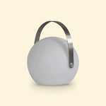 Lampe ronde d’extérieur solaire éteinte, avec anse métallique arquée, posée sur un fond beige clair.