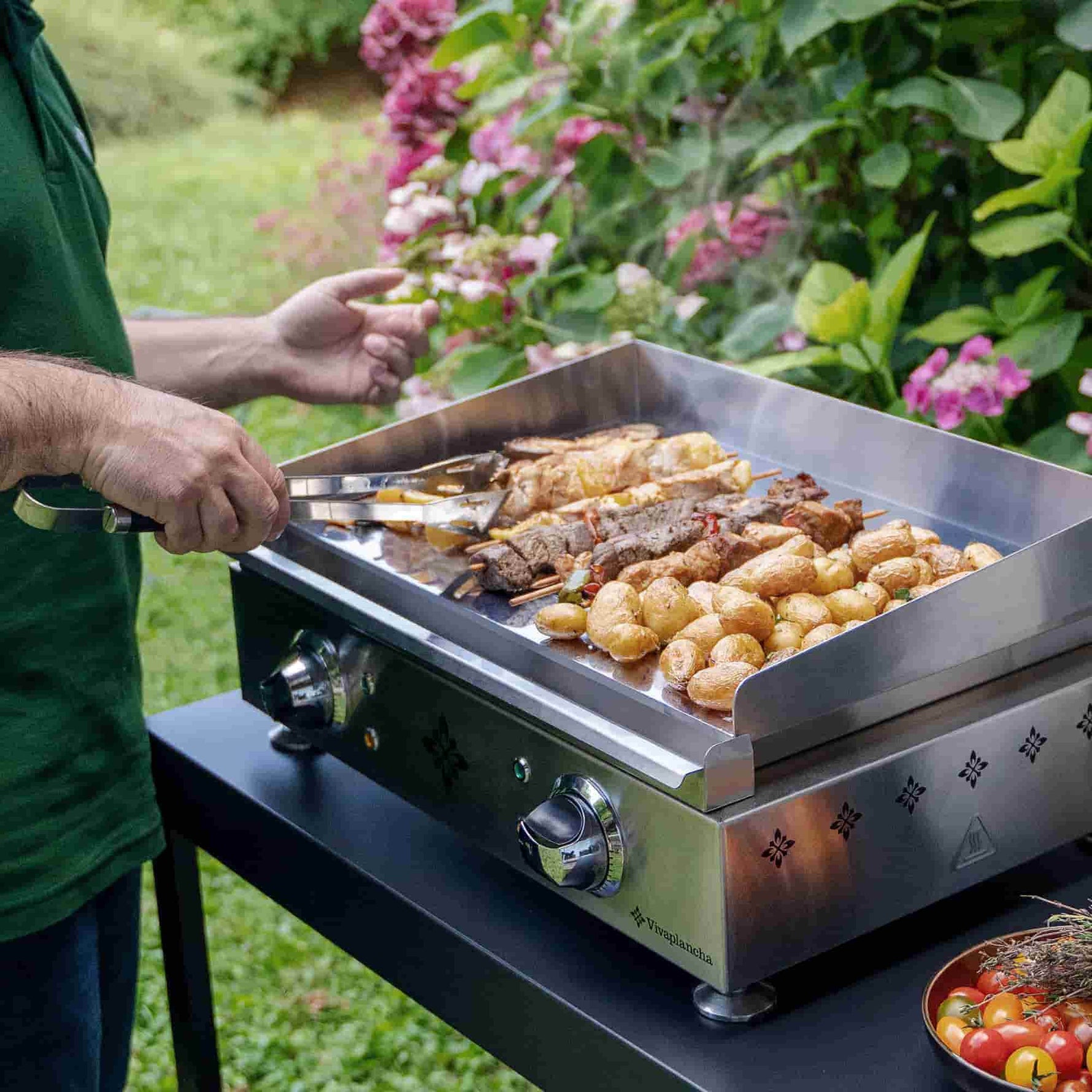 Plancha Vivaplancha en inox en fonctionnement, avec pommes de terre et brochettes grillées, photographiée en extérieur avec des personnes floues en arrière-plan.