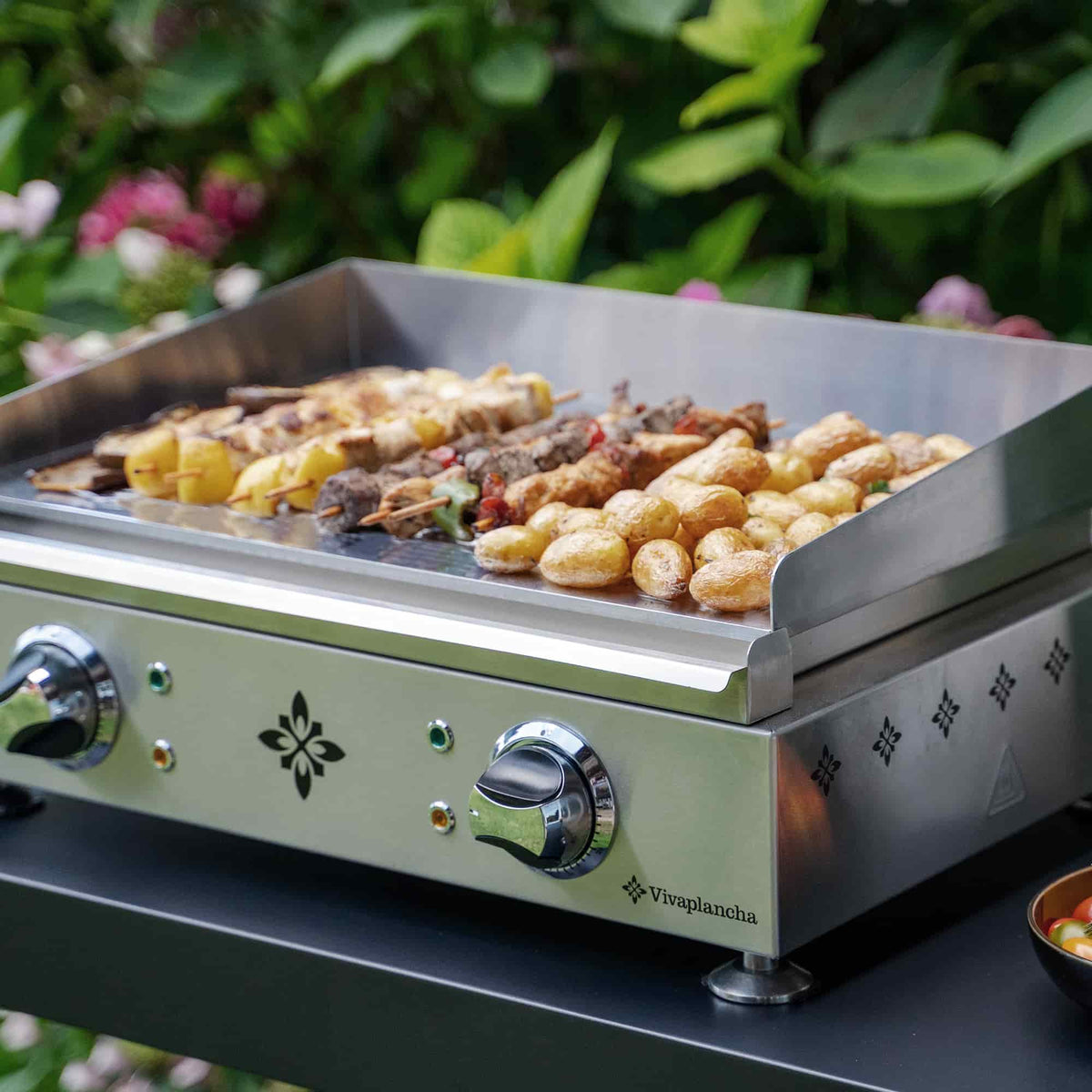 Plancha électrique Vivaplancha en inox avec plaque en chrome dur en pleine utilisation, faisant griller brochettes et pommes de terre, dans un décor extérieur verdoyant.
