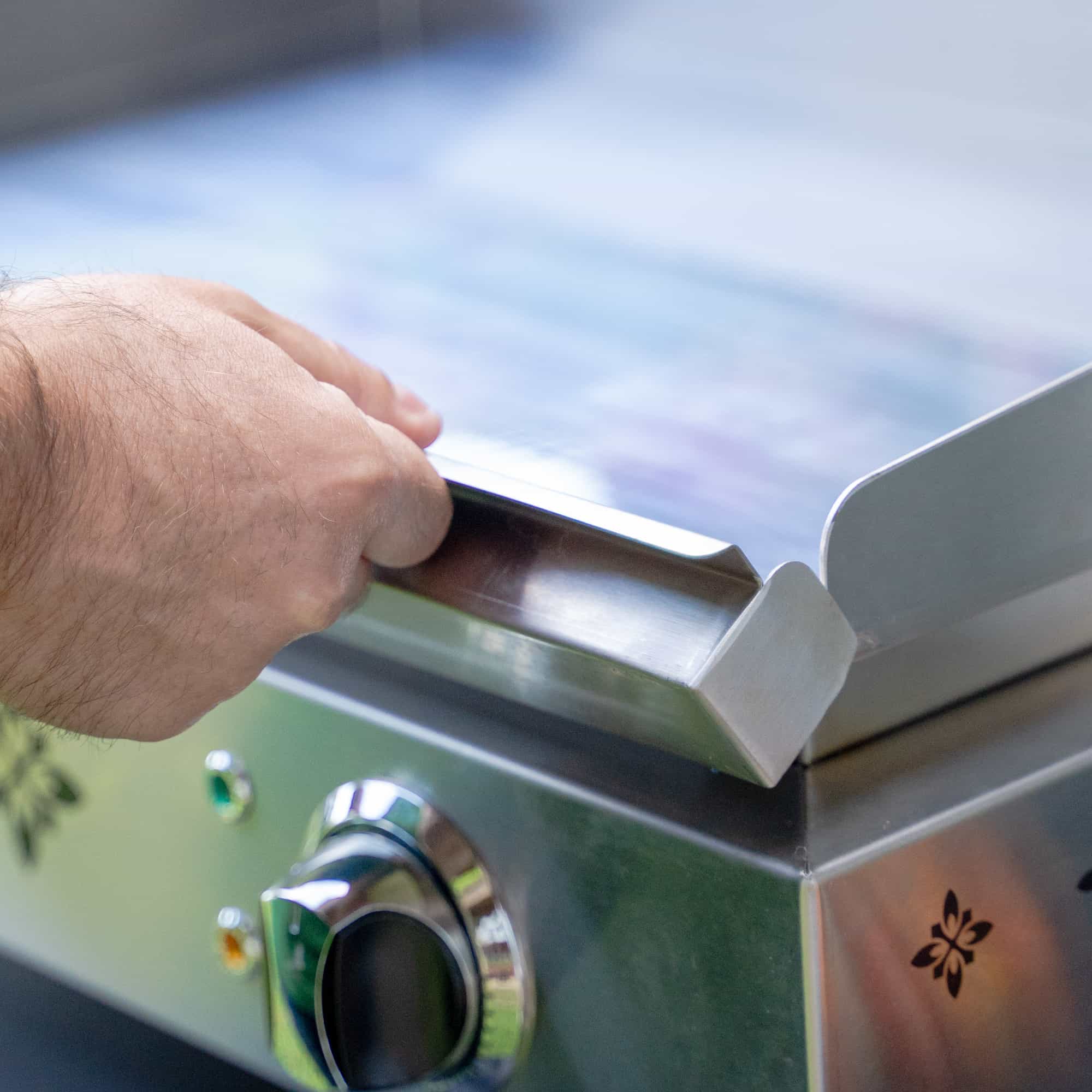 Nahaufnahme der Hand einer Person, die den Rand der Edelstahlplatte einer elektrischen Vivaplancha-Plancha bedient, mit dem Einstellknopf im Vordergrund sichtbar.
