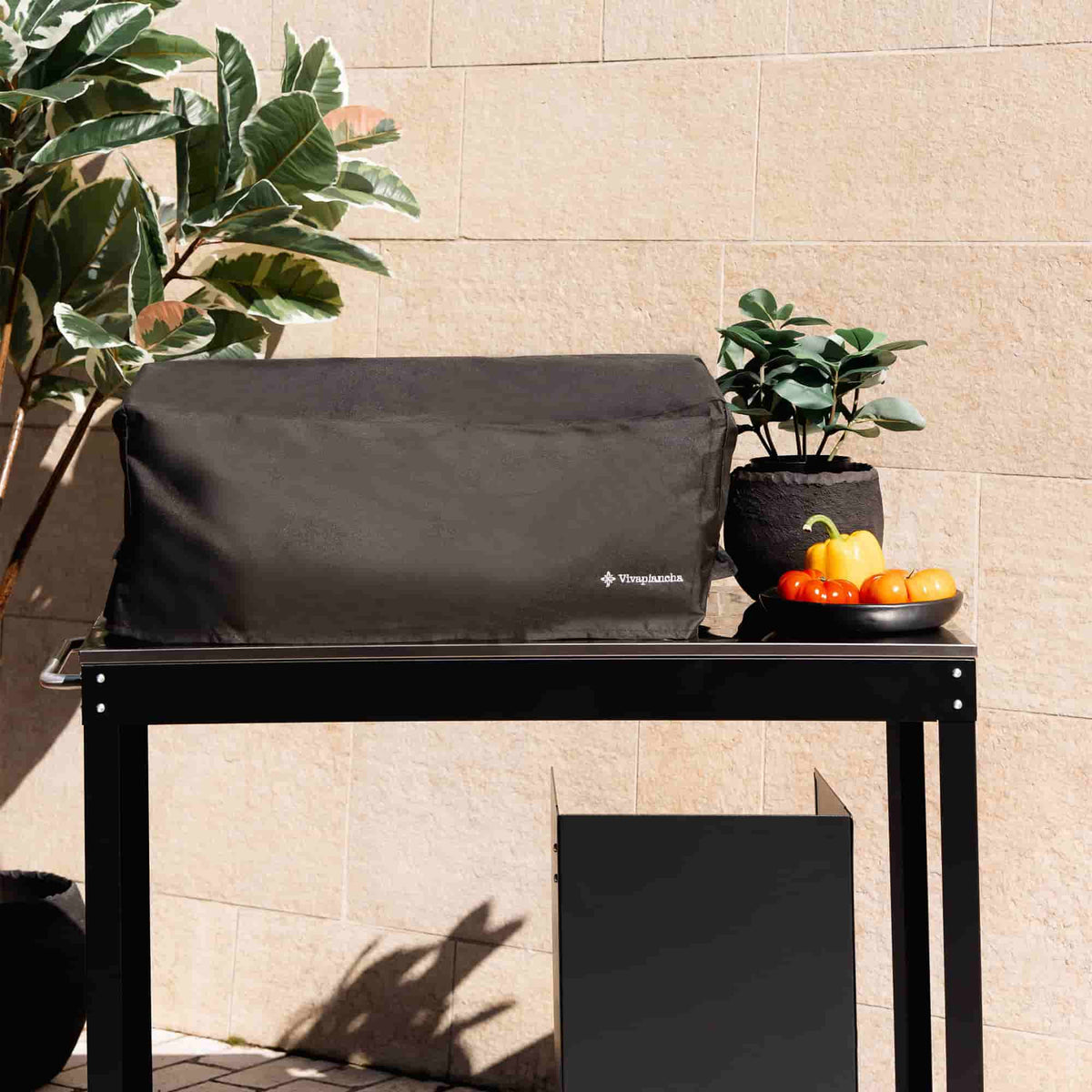 Housse noire de protection pour plancha deux brûleurs Vivaplancha, posée sur une desserte en extérieur, entourée de plantes et de légumes, présentée en situation réelle.