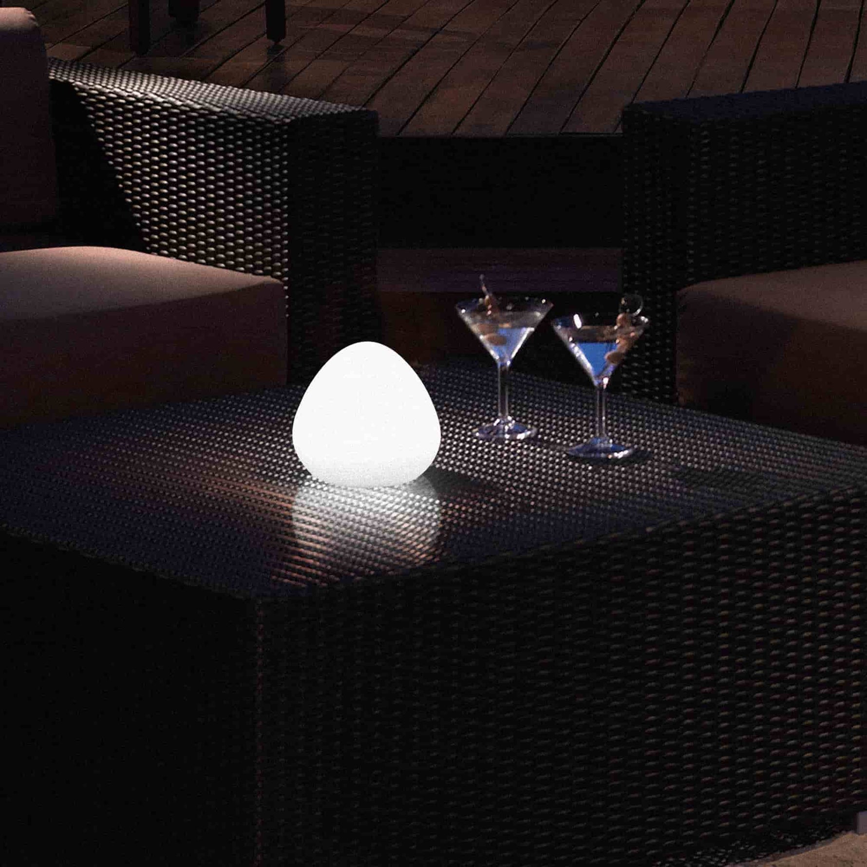 Lampe solaire d’extérieur en forme de noisette, posée sur une table en résine tressée, diffusant une lumière blanche douce dans une ambiance de terrasse nocturne.