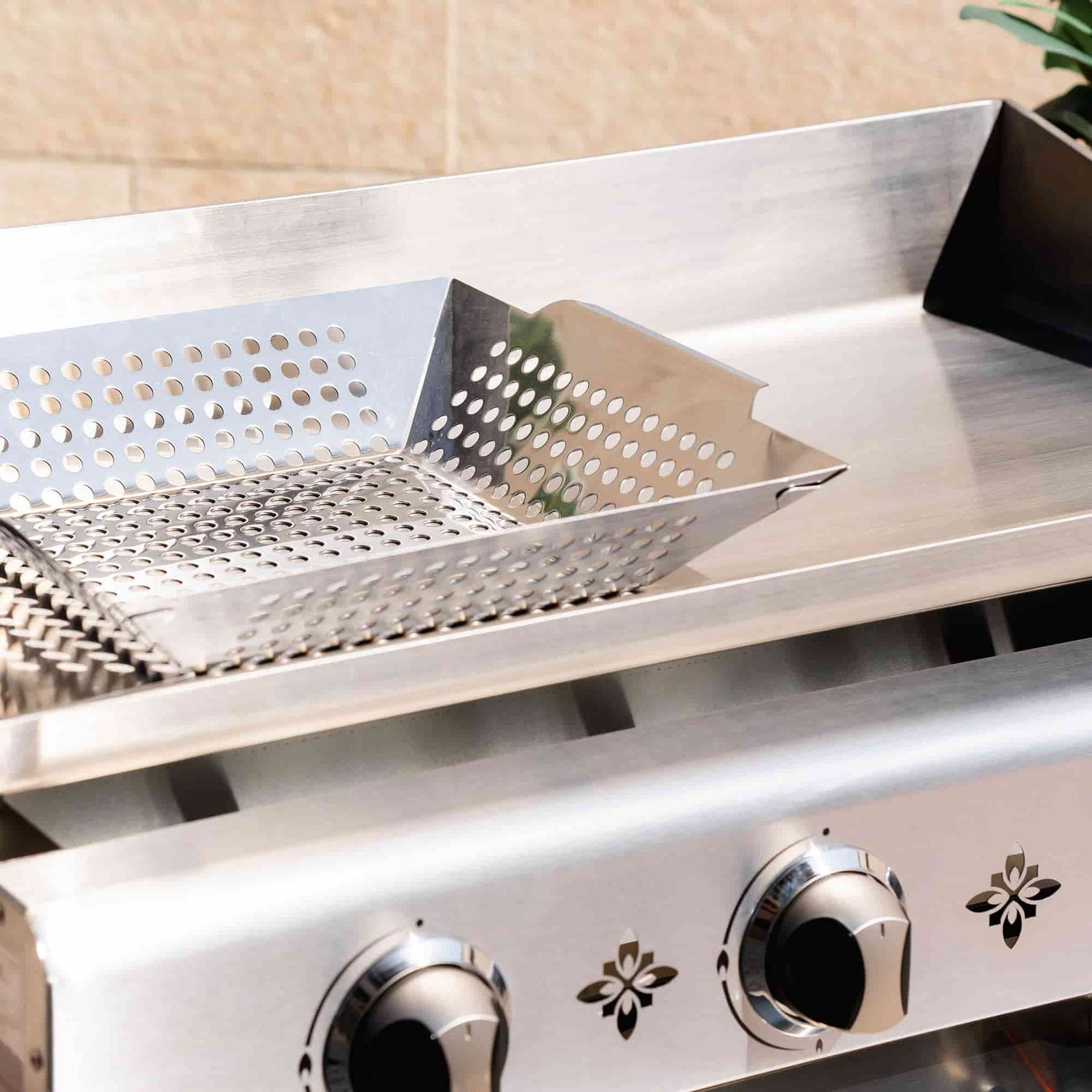 Panier de cuisson perforé en inox posé sur la plaque d’une plancha inox à trois brûleurs, présenté en gros plan en extérieur.
