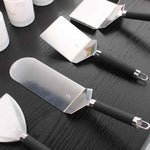 Ensemble de spatules en inox avec manches noirs, disposées sur un plan de travail en bois foncé, montrées en vue rapprochée.