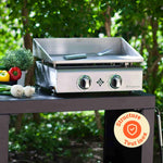 Edelstahl-Plancha Vivaplancha auf einem Außenwagen, begleitet von frischem Gemüse, mit einem „Komplett-Edelstahl“-Abzeichen unten rechts.