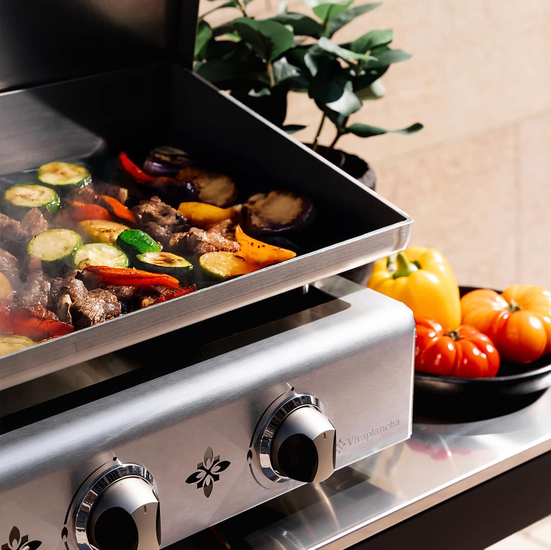 Plancha gaz Vivaplancha Pimientos en inox en pleine cuisson, avec légumes grillés colorés sur la plaque et boutons de contrôle en métal en premier plan, décorée de tomates et d’un poivron sur le côté.