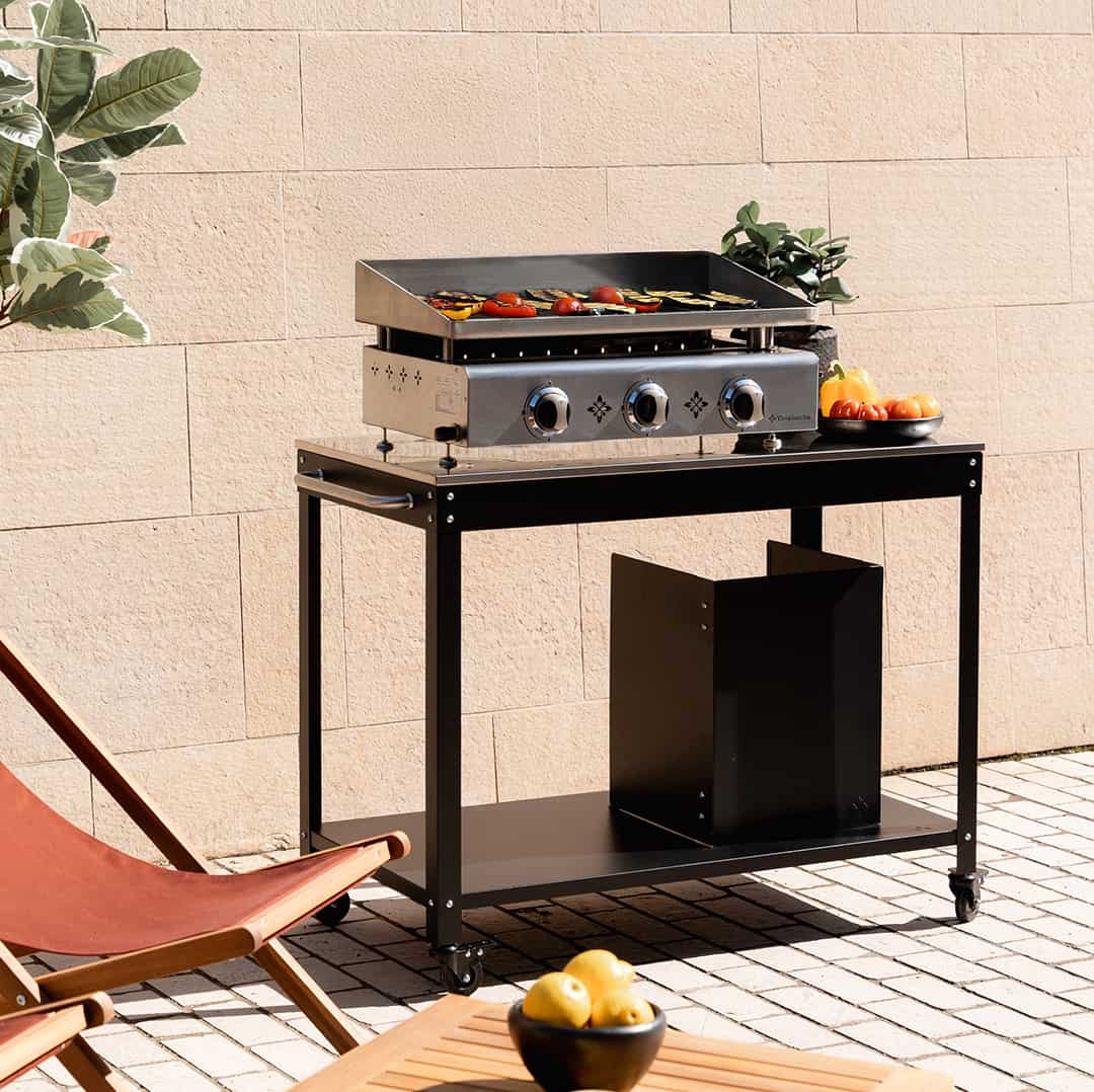 Plancha gaz Vivaplancha installée sur une desserte noire en extérieur, avec légumes en cuisson sur la plaque, bol de tomates et poivrons sur le côté, et transat en premier plan.